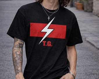 May include: Black t-shirt with a red rectangle and a white lightning bolt graphic. The text "T.G." is below the lightning bolt. The shirt is worn with black jeans and silver jewelry.