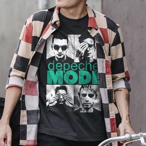 Può includere: Una t-shirt nera a maniche corte con una stampa grafica della band Depeche Mode. Il design include quattro ritratti in bianco e nero dei membri della band con il testo "depeche MODE" in verde, indossata sotto una camicia a quadri rossa, nera e beige.