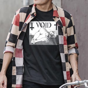May include: A black t-shirt featuring a white rectangular graphic print. The graphic depicts a reclining figure with an open mouth, the text "VOID" in a stylised font, and two inverted crosses on either side, worn under a red, black, and beige plaid shirt.