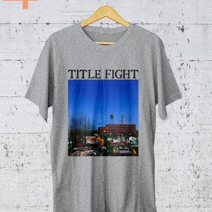 May include: Gray t-shirt with the text "TITLE FIGHT" printed on the front. The shirt features a graphic of a building with a blue sky in the background.