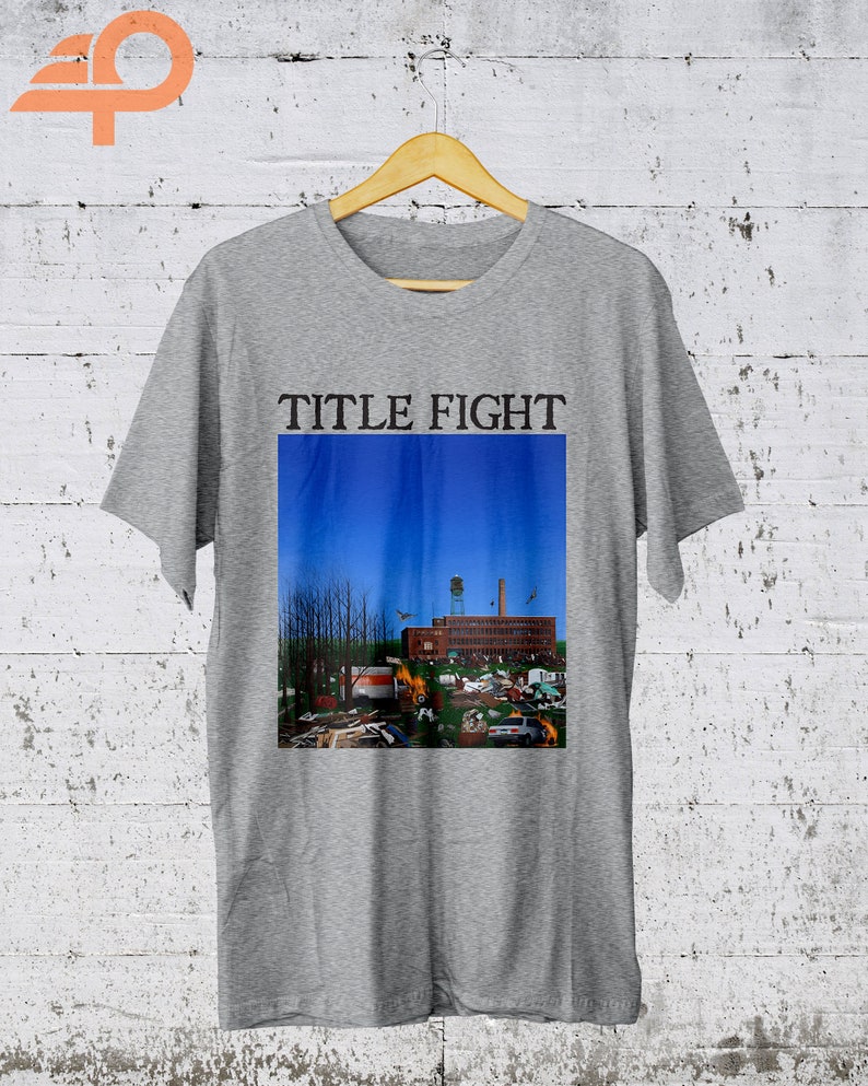 May include: Gray t-shirt with the text "TITLE FIGHT" printed on the front. The shirt features a graphic of a building with a blue sky in the background.