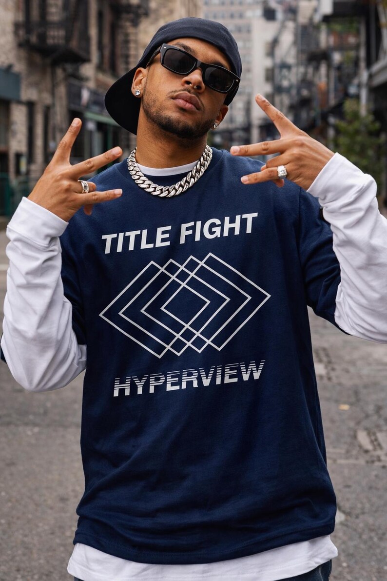 May include: Navy blue long-sleeve shirt with the text "TITLE FIGHT" and "HYPERVIEW" in white. The shirt features a geometric design. The model is wearing a black cap, sunglasses, and a silver chain.