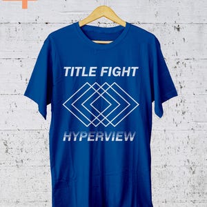 K&ouml;nnte beinhalten: Ein k&ouml;nigsblaues T-Shirt mit den Worten "TITLE FIGHT" &uuml;ber einem wei&szlig;en geometrischen Design und dem Wort "HYPERVIEW" darunter. Das T-Shirt h&auml;ngt an einem Holzb&uuml;gel vor einer wei&szlig;en Backsteinmauer.