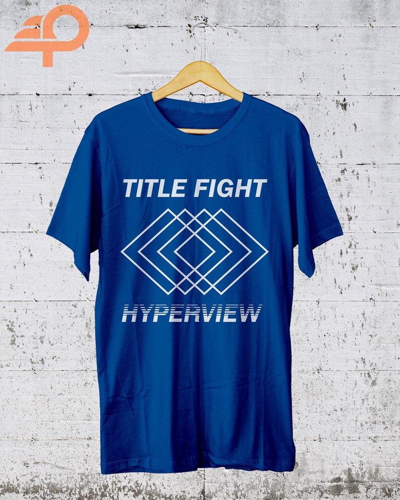K&ouml;nnte beinhalten: Ein k&ouml;nigsblaues T-Shirt mit den Worten "TITLE FIGHT" &uuml;ber einem wei&szlig;en geometrischen Design und dem Wort "HYPERVIEW" darunter. Das T-Shirt h&auml;ngt an einem Holzb&uuml;gel vor einer wei&szlig;en Backsteinmauer.