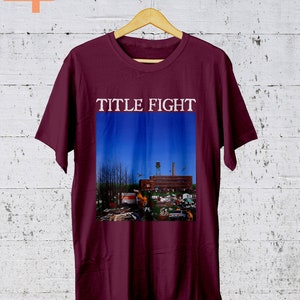 May include: A maroon t-shirt with the band name "TITLE FIGHT" printed in white on the front. The shirt features a graphic of a building with a blue sky in the background.