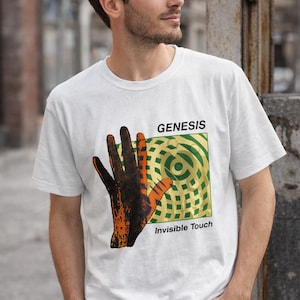 May include: White t-shirt with the band name "Genesis" and the album title "Invisible Touch." The design features a hand in orange and black, set against a green and gold background. The shirt has a crew neck.
