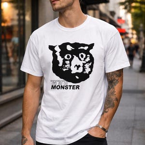 May include: White t-shirt featuring a black graphic of a monster face with the text "R.E.M. MONSTER" in black. The shirt is worn by a person in black pants.