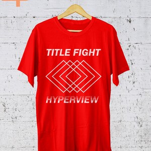 K&ouml;nnte beinhalten: Ein leuchtend rotes T-Shirt mit dem wei&szlig;en Schriftzug "TITLE FIGHT" &uuml;ber einem geometrischen Rautenmuster. Darunter steht das Wort "HYPERVIEW" in einer verzerrten Schriftart. Das T-Shirt h&auml;ngt an einem Holzkleiderb&uuml;gel.