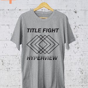K&ouml;nnte beinhalten: Hellgraues T-Shirt mit dem Schriftzug "TITLE FIGHT" &uuml;ber einem geometrischen Rautenmuster und dem Wort "HYPERVIEW" darunter. Das T-Shirt h&auml;ngt an einem Holzb&uuml;gel vor einer wei&szlig;en Backsteinmauer.