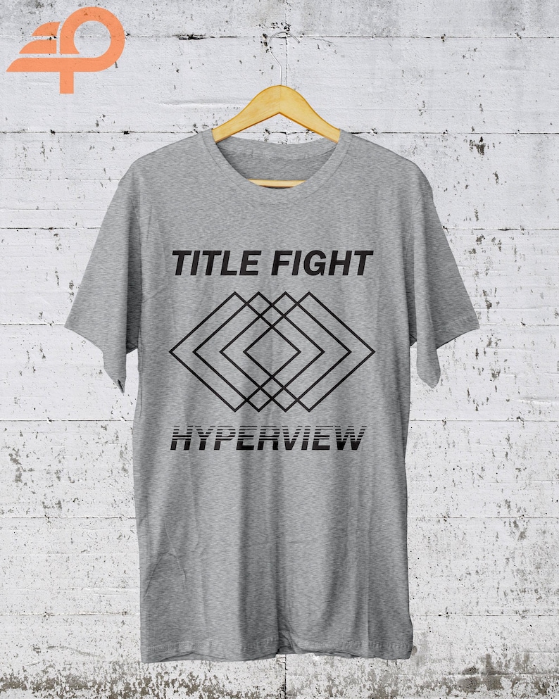 K&ouml;nnte beinhalten: Hellgraues T-Shirt mit dem Schriftzug "TITLE FIGHT" &uuml;ber einem geometrischen Rautenmuster und dem Wort "HYPERVIEW" darunter. Das T-Shirt h&auml;ngt an einem Holzb&uuml;gel vor einer wei&szlig;en Backsteinmauer.