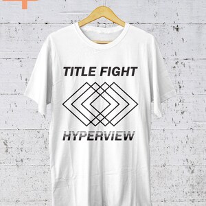 K&ouml;nnte beinhalten: Wei&szlig;es T-Shirt mit dem Text "TITLE FIGHT" &uuml;ber einem geometrischen Rautenmuster und dem Wort "HYPERVIEW" darunter. Das T-Shirt h&auml;ngt an einem Holzb&uuml;gel vor einer wei&szlig;-grauen Wand.