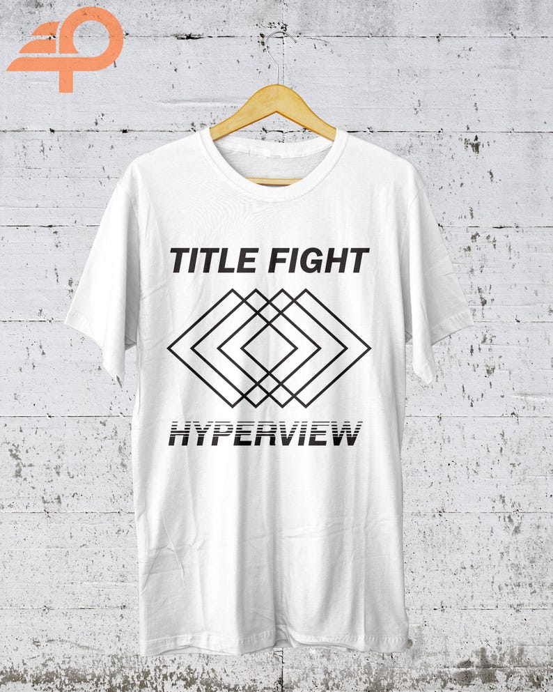 K&ouml;nnte beinhalten: Wei&szlig;es T-Shirt mit dem Text "TITLE FIGHT" &uuml;ber einem geometrischen Rautenmuster und dem Wort "HYPERVIEW" darunter. Das T-Shirt h&auml;ngt an einem Holzb&uuml;gel vor einer wei&szlig;-grauen Wand.