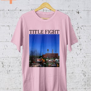 May include: Pink t-shirt with a graphic print of a building with a blue sky and the text "TITLE FIGHT" above the image.
