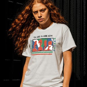 May include: A white t-shirt with the words "TALKIN HEADS" and a colourful graphic design. The graphic features red, green, and blue shapes. The shirt is worn by a person with long, red hair.