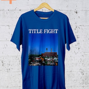 May include: A royal blue t-shirt with the words "TITLE FIGHT" printed in white on the front. The shirt features a graphic of a building with a blue sky and a fire in the foreground.
