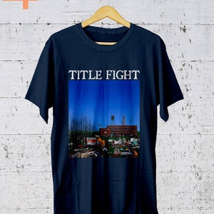 May include: A navy blue t-shirt with a graphic of a building with a blue sky and the text "TITLE FIGHT" printed in white.