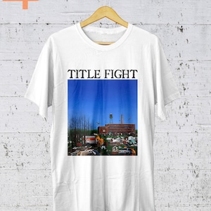 May include: White t-shirt with a blue and red graphic of a building with a blue sky and the text "TITLE FIGHT" printed in black.