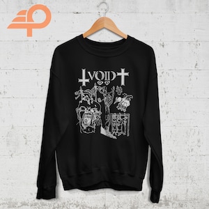 May include: Black sweatshirt with a white graphic design. The design includes the word "VOID" with a cross, and several illustrations of figures. The sweatshirt is hanging on a wooden hanger.