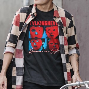 May include: Black t-shirt with a graphic of four faces in red and blue, with the text "TALKING HEADS" above and "Remain in Light" below. The person is wearing a checkered red, black, and beige button-down shirt.