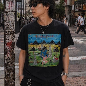 May include: Black Talking Heads t-shirt with a colourful graphic of a landscape with figures and buildings. The shirt is worn by a person wearing sunglasses and a silver necklace, in an urban setting.