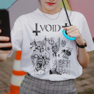 Może przedstawiać: Biały t-shirt z krótkim rękawem z czarnym nadrukiem graficznym na klatce piersiowej. Wzór zawiera słowo VOID stylizowaną czcionką otoczone dwoma krzyżami, nad szczegółową ilustracją krzyczącej twarzy oraz różnymi postaciami.