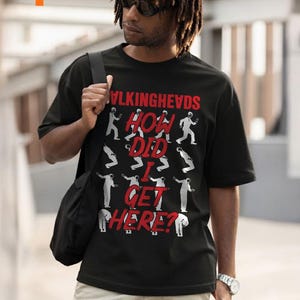 May include: Black t-shirt with the text "VLKINGHEVDS HOW DID I GET HERE?" in red and white. The shirt features a graphic of stylized figures. The person in the image is wearing sunglasses and a watch.
