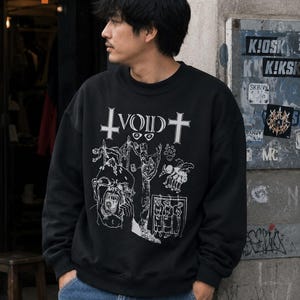 May include: A black crewneck sweatshirt featuring a white graphic print on the chest. The design includes the text "VOID" flanked by two crosses, along with detailed illustrations of a screaming face, figures, and various abstract symbols.