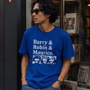 Puede incluir: Una camiseta azul real con el texto "Barry & Robin & Maurice" y una imagen en blanco y negro de tres hombres. La camiseta la lleva una persona con gafas de sol y vaqueros azules.