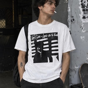 Può includere: Una t-shirt bianca a maniche corte con una stampa grafica in bianco e nero sul petto. La grafica mostra una silhouette su strisce orizzontali con il testo "The Cure - Let's Go To Bed" stampato in stile manoscritto sopra l'immagine.