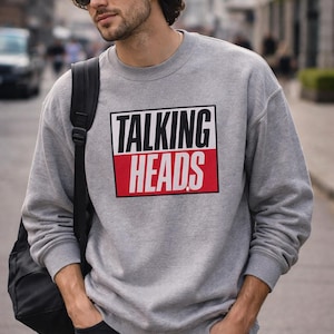 May include: A grey sweatshirt featuring the text "TALKING HEADS" in a black and red rectangular design. The sweatshirt is worn by a person wearing sunglasses and carrying a black backpack.