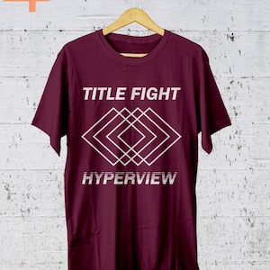 K&ouml;nnte beinhalten: Ein weinrotes T-Shirt mit dem Schriftzug "TITLE FIGHT" &uuml;ber einem wei&szlig;en geometrischen Design und dem Wort "HYPERVIEW" darunter. Das T-Shirt h&auml;ngt an einem Holzb&uuml;gel vor einer wei&szlig; get&uuml;nchten Wand.