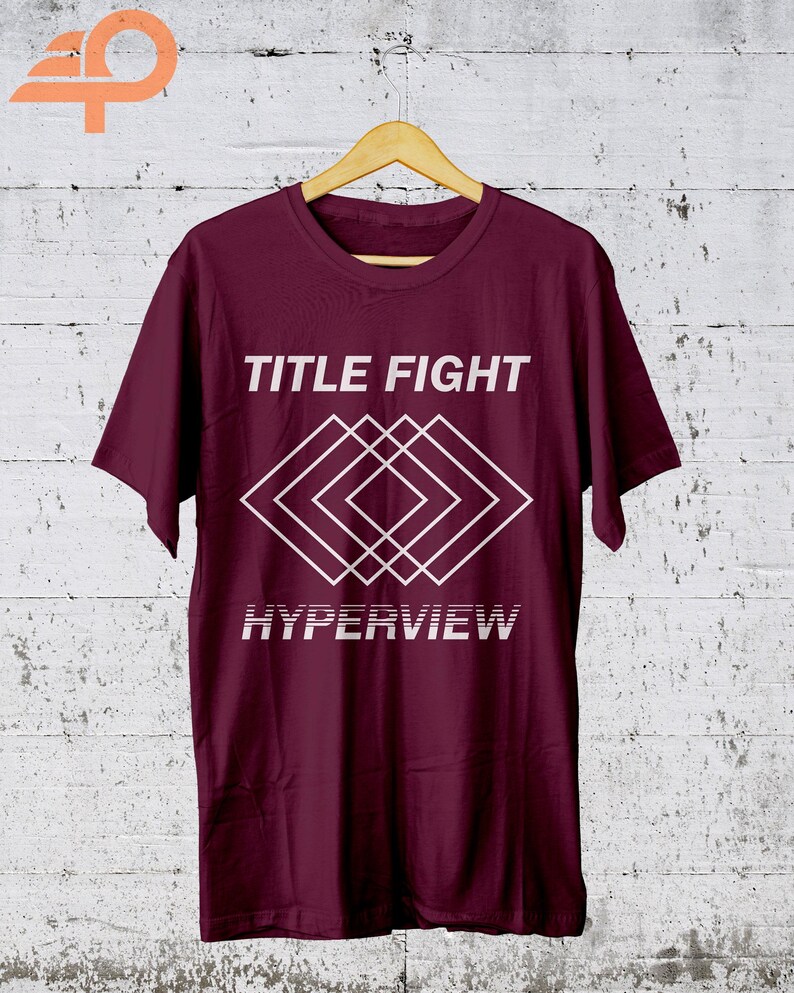 K&ouml;nnte beinhalten: Ein weinrotes T-Shirt mit dem Schriftzug "TITLE FIGHT" &uuml;ber einem wei&szlig;en geometrischen Design und dem Wort "HYPERVIEW" darunter. Das T-Shirt h&auml;ngt an einem Holzb&uuml;gel vor einer wei&szlig; get&uuml;nchten Wand.