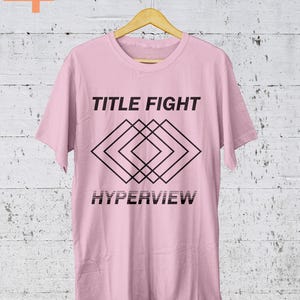 K&ouml;nnte beinhalten: Ein hellrosa T-Shirt mit den Worten "TITLE FIGHT" &uuml;ber einem schwarzen geometrischen Design und dem Wort "HYPERVIEW" darunter. Das T-Shirt h&auml;ngt an einem Holzb&uuml;gel vor einem wei&szlig;en und grauen Hintergrund.