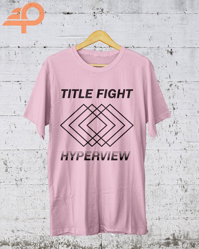 K&ouml;nnte beinhalten: Ein hellrosa T-Shirt mit den Worten "TITLE FIGHT" &uuml;ber einem schwarzen geometrischen Design und dem Wort "HYPERVIEW" darunter. Das T-Shirt h&auml;ngt an einem Holzb&uuml;gel vor einem wei&szlig;en und grauen Hintergrund.