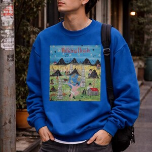 May include: A vibrant blue sweatshirt with a graphic print. The print features the text "Talking Heads" and a landscape illustration with mountains, buildings, and figures. The sweatshirt is a solid colour and has a crew neck.