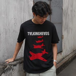 May include: Black t-shirt featuring the white text "TALKINGHEADS" and a series of red airplane graphics. The person in the image is wearing light grey trousers and a silver watch, standing by a concrete wall.