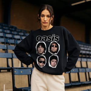 May include: Black sweatshirt featuring the band name "oasis" in white, with four band member portraits. The model is wearing beige sweatpants. The background shows blue stadium seating.