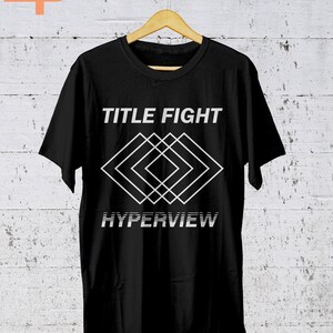 K&ouml;nnte beinhalten: Schwarzes T-Shirt mit dem Schriftzug "TITLE FIGHT" &uuml;ber einem wei&szlig;en geometrischen Design und dem Wort "HYPERVIEW" darunter. Das T-Shirt h&auml;ngt an einem Holzb&uuml;gel vor einer wei&szlig;en Backsteinmauer.