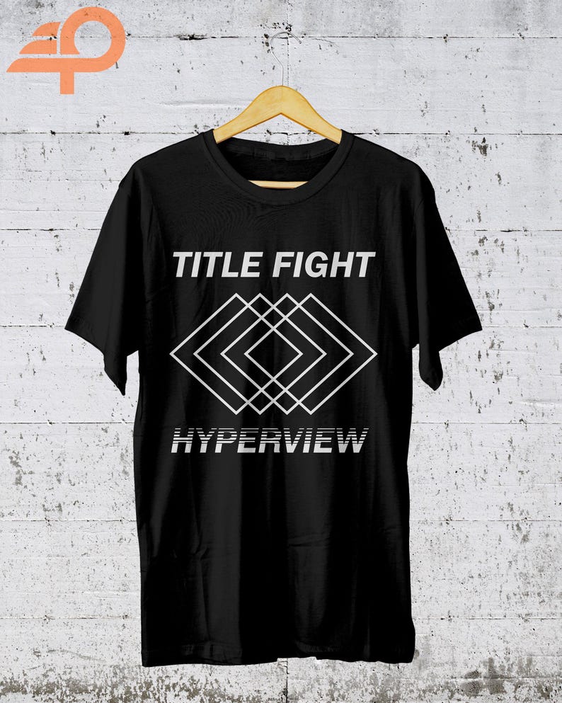 K&ouml;nnte beinhalten: Schwarzes T-Shirt mit dem Schriftzug "TITLE FIGHT" &uuml;ber einem wei&szlig;en geometrischen Design und dem Wort "HYPERVIEW" darunter. Das T-Shirt h&auml;ngt an einem Holzb&uuml;gel vor einer wei&szlig;en Backsteinmauer.