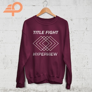 May include: A maroon sweatshirt with a white graphic design. The graphic features a geometric pattern of overlapping squares and the text "TITLE FIGHT" and "HYPERVIEW".