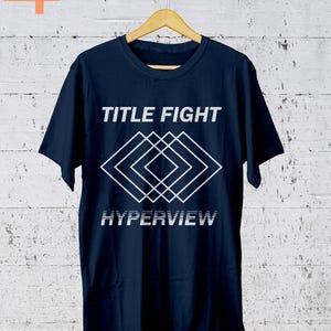 K&ouml;nnte beinhalten: Marineblaues T-Shirt mit dem Schriftzug "TITLE FIGHT" &uuml;ber einem wei&szlig;en geometrischen Design und dem Wort "HYPERVIEW" darunter. Das T-Shirt h&auml;ngt an einem Holzb&uuml;gel vor einem wei&szlig;-grauen Hintergrund.