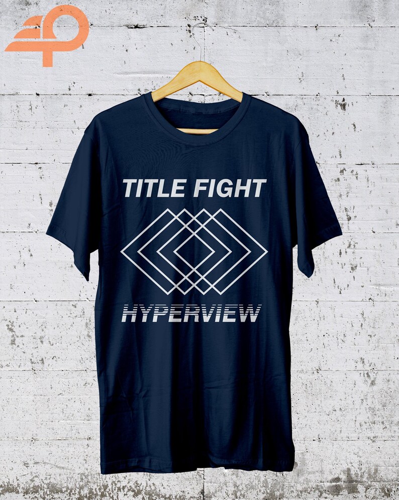 K&ouml;nnte beinhalten: Marineblaues T-Shirt mit dem Schriftzug "TITLE FIGHT" &uuml;ber einem wei&szlig;en geometrischen Design und dem Wort "HYPERVIEW" darunter. Das T-Shirt h&auml;ngt an einem Holzb&uuml;gel vor einem wei&szlig;-grauen Hintergrund.