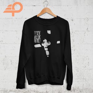 May include: Black sweatshirt featuring a white graphic of a woman's face and the band name "Siouxsie and the Banshees". The design is a stylized, minimalist portrait. The sweatshirt is hanging on a wooden hanger.