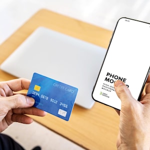 May include: A person holding a blue credit card and a smartphone with a blank screen. The phone displays the text "PHONE MOCKUP" and other text. A laptop is visible in the background on a wooden surface.