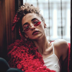 May include: A woman wearing red oval sunglasses, a red scarf with white polka dots, and a red fluffy jacket. She is also wearing a white tank top and large silver hoop earrings. Her hair is styled in a bun.