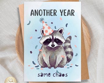 May include: A rectangular greeting card featuring a watercolor illustration of a raccoon wearing a peach-colored party hat with white polka dots. The text reads "ANOTHER YEAR" at the top and "same chaos" at the bottom, surrounded by confetti.