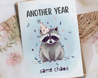 May include: Greeting card with a raccoon wearing a party hat, surrounded by confetti. The card reads "ANOTHER YEAR" above the raccoon and "same chaos" below. The card is on a white surface with a brown envelope.
