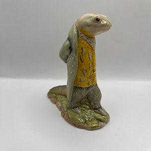 May include: A ceramic figurine of a lizard wearing a grey coat, yellow shirt, and grey trousers. The lizard is standing on a green base.