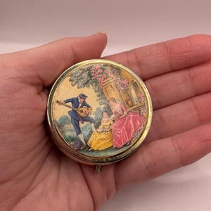 May include: A round, gold-toned metal compact with a hinged lid. The lid features a colourful painted scene of a man playing a lute for two women in a garden setting.