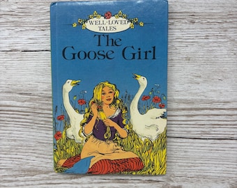 May include: A blue hardcover book titled Well-Loved Tales The Goose Girl. The cover features an illustration of a person with long blonde hair sitting in a field of red poppies between two white geese, with a small ladybug above the title text.
